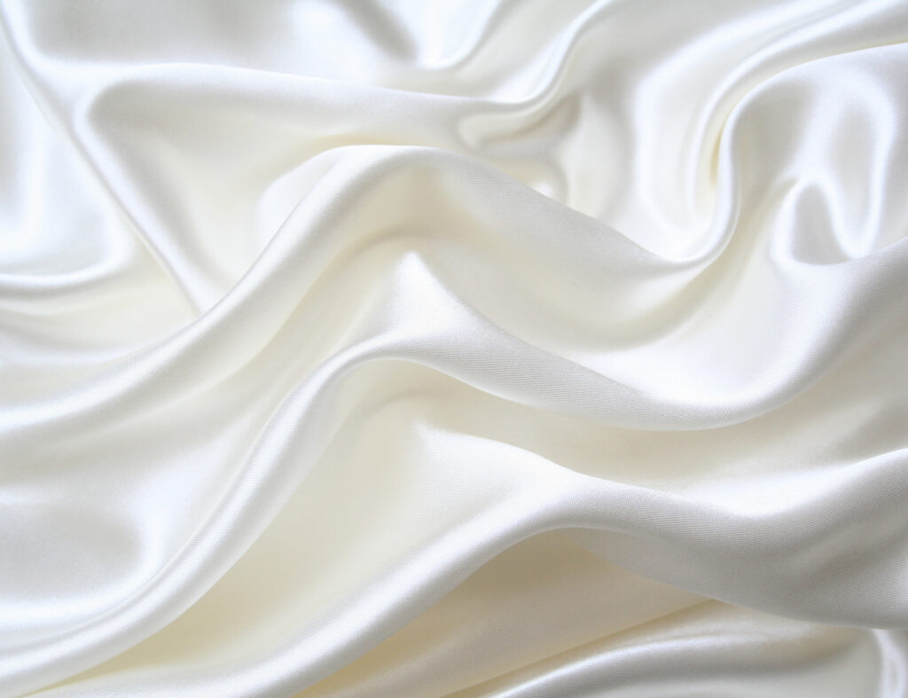 Close-up of luxurious white silk fabric draped in elegant flowing folds with soft sheen, pearl highlights, and gentle shadows creating a serene, minimalist aesthetic.