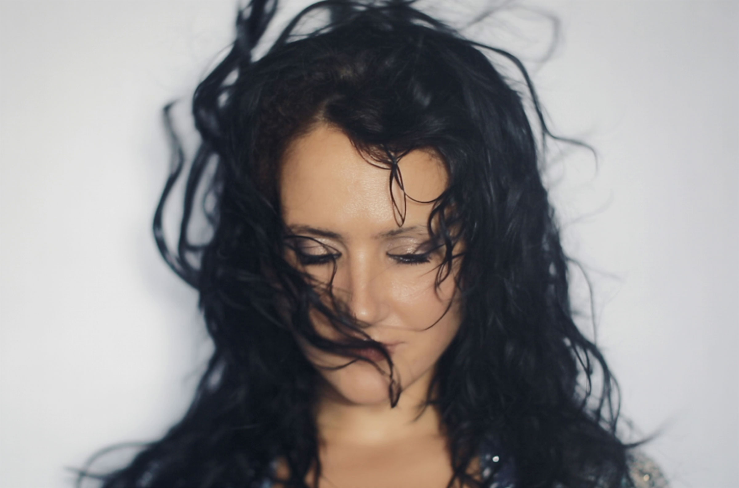 Artistic close-up portrait of a woman with wind-swept black hair flowing across her face against a stark white background, eyes closed in contemplative serenity.