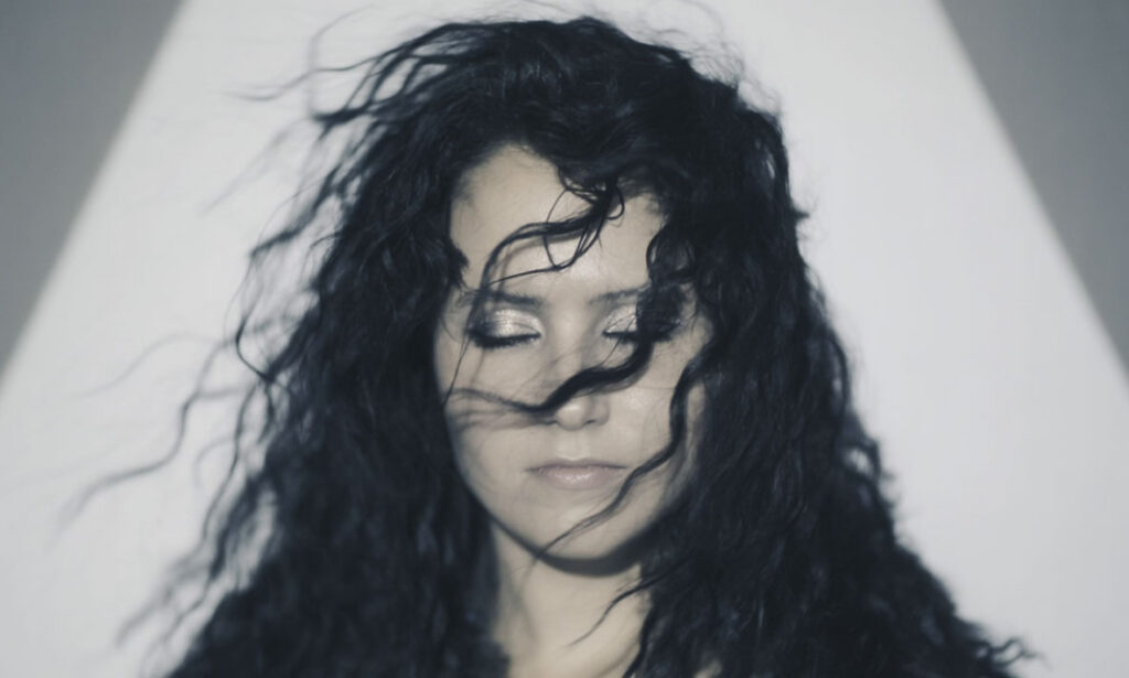 Fine art editorial portrait of a woman with flowing black hair swept across her face, eyes closed, set against a clean white background with dramatic contrast.