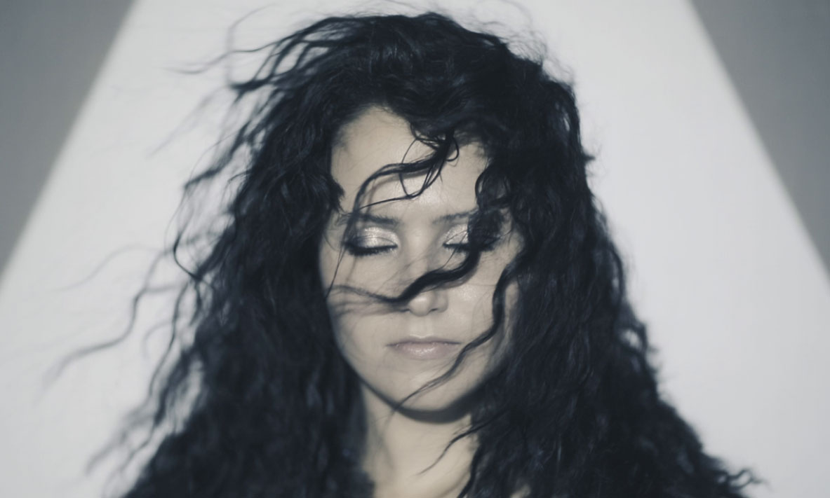 Fine art editorial portrait of a woman with flowing black hair swept across her face, eyes closed, set against a clean white background with dramatic contrast.